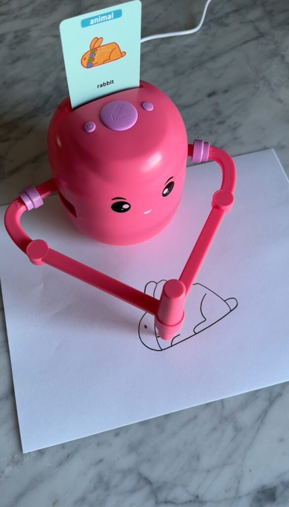 A pink educational robot with cute facial features uses its drawn arms to sketch a rabbit on white paper. A card labeled "animal" and "rabbit" is inserted into the robot. The scene is set on a marble surface.