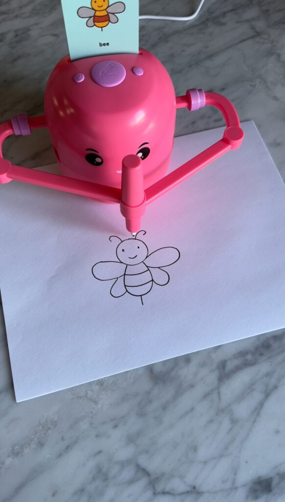 A pink drawing robot for kids draws a simple, smiling bee on white paper set on a gray marble surface. A small card with a bee illustration is clipped to the robot.