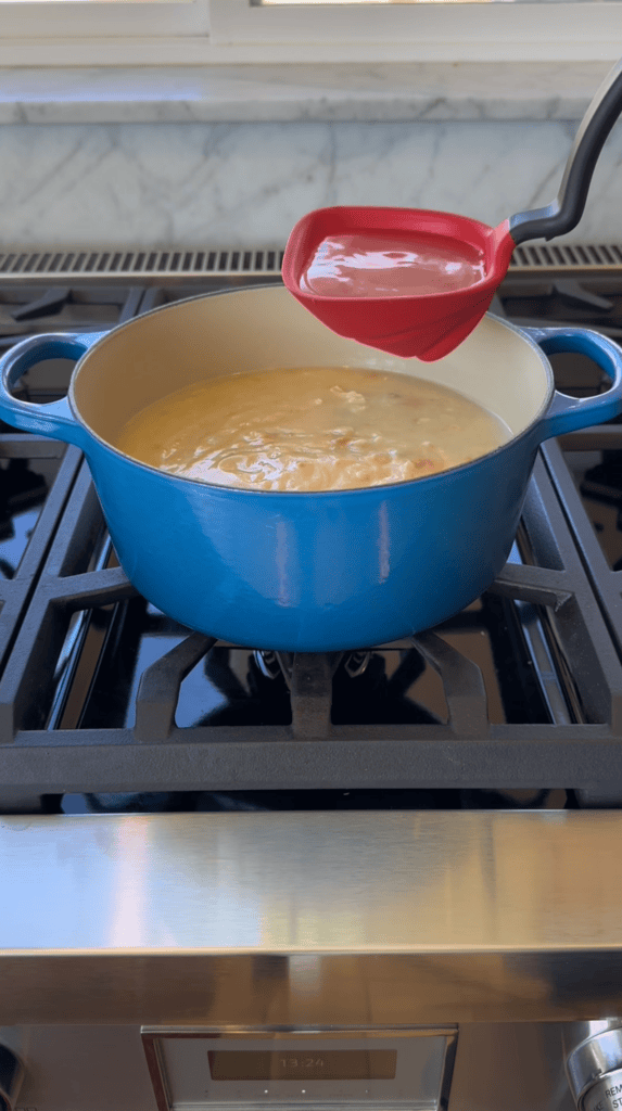 A blue Dutch oven sits on a stovetop, filled with soup. A Fold Flat Ladle is held above the pot, showing a serving of the creamy soup inside. The stove and part of a marble backsplash are visible in this cozy kitchen scene.