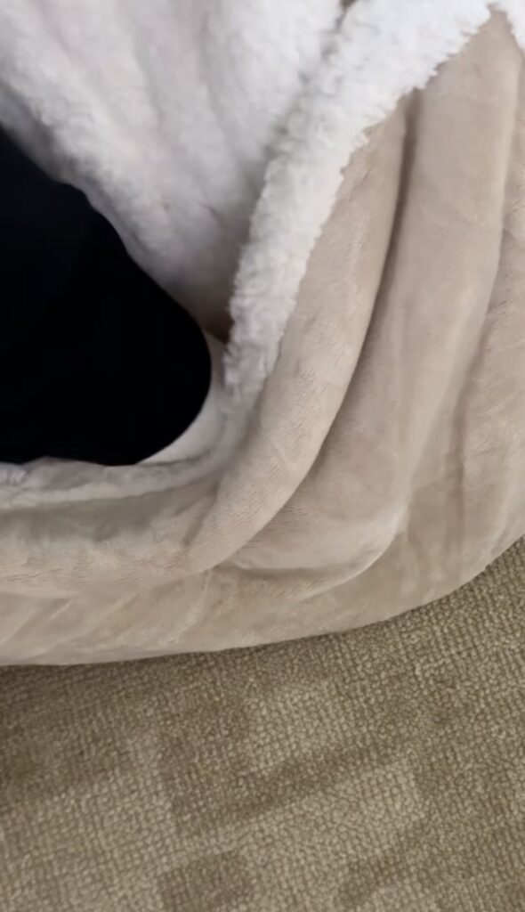 A close-up of a cozy blanket with a white fluffy lining draped over the edge of a beige carpet, perfect for moms seeking comfort. A small section of black fabric, possibly from a wearable heated blanket, peeks out from under the blanket.