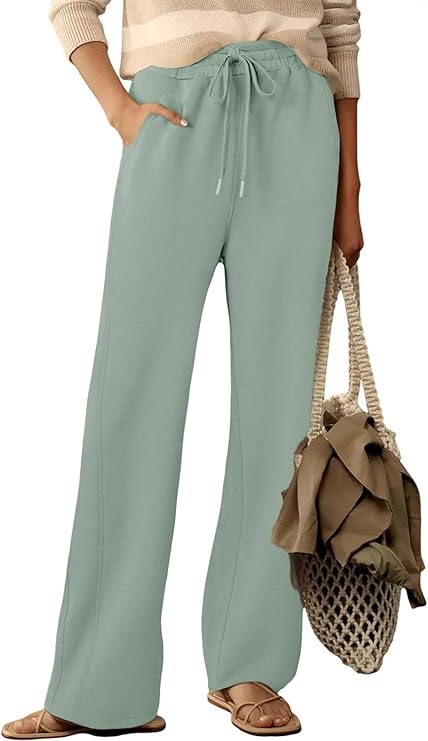 A person wearing loose, light green drawstring pants, a beige and white striped sweater, beige sandals, and holding a netted tote bag with a fabric item inside. Only the lower body is visible.