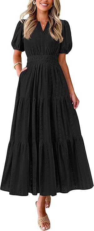 A woman wearing a black, short-sleeve, ankle-length maxi dress with a fitted waist and tiered skirt. She has one hand in her pocket and is wearing sandals and hoop earrings.