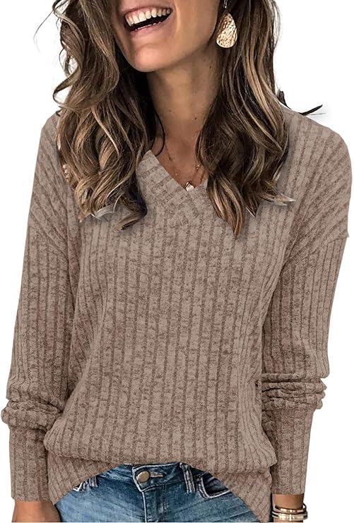 A woman with wavy brown hair wears a beige, ribbed, V-neck sweater with long sleeves and blue jeans. She is smiling and accessorized with large earrings.