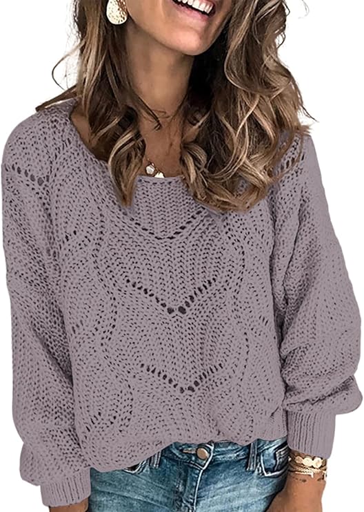 A woman wearing a light purple, loose-knit sweater with a wavy pattern, blue jeans, dangle earrings, and layered necklaces, smiling with wavy hair over her shoulders.