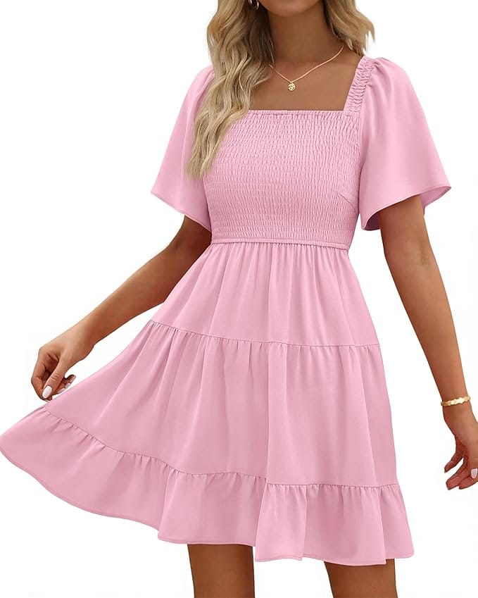 A woman wearing a light pink, short-sleeve, tiered dress with a smocked bodice and square neckline. She holds out one side of the skirt, and her long, wavy hair is blonde. She wears a gold necklace and bracelet.