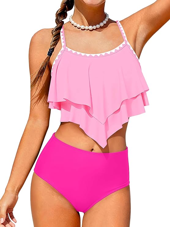 A woman wearing a light pink ruffled tankini top and high-waisted hot pink bikini bottoms, accessorized with a beaded necklace and a side braid, smiles with one arm raised.