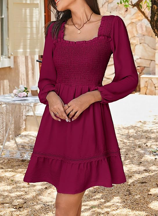 A woman wearing a long-sleeved, square-neck, burgundy dress with a smocked bodice and flared skirt stands outdoors near a stone wall and a white table with flowers.