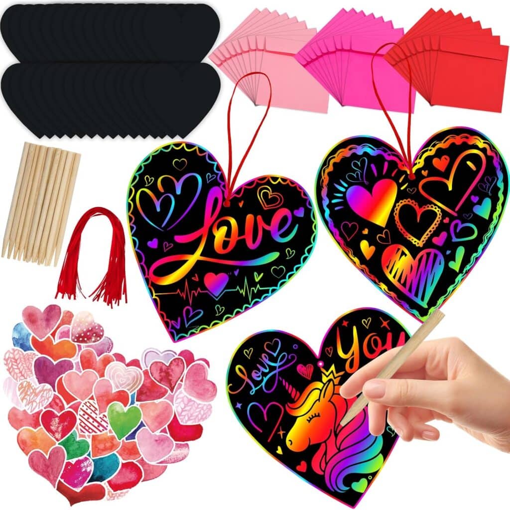 A colorful scratch art kit featuring heart-shaped scratch cards, colored envelopes, black scratch boards, wooden styluses, red strings, and a hand drawing a rainbow design on a heart-shaped scratch card.