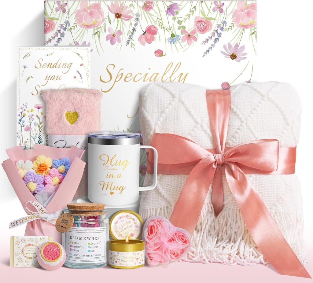 A gift box with a floral design contains a white blanket tied with a pink ribbon, a mug labeled "Hug in a Mug," a pink fuzzy sock, colorful flower-shaped soaps, candles, a card, and other self-care items.