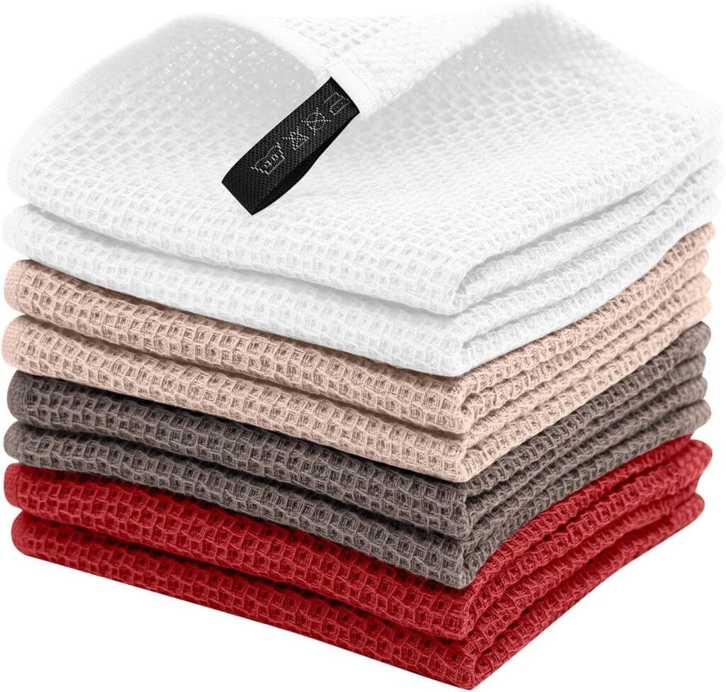 A stack of eight waffle-textured kitchen towels in white, beige, light brown, brown, and red shades. The top towel has a black tag attached to one corner.