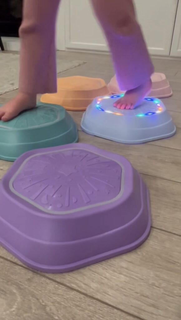 A child steps barefoot on colorful sensory balance toys, including a purple stepping stone in the foreground and a blue one with twinkling lights, all arranged on a wooden floor indoors.