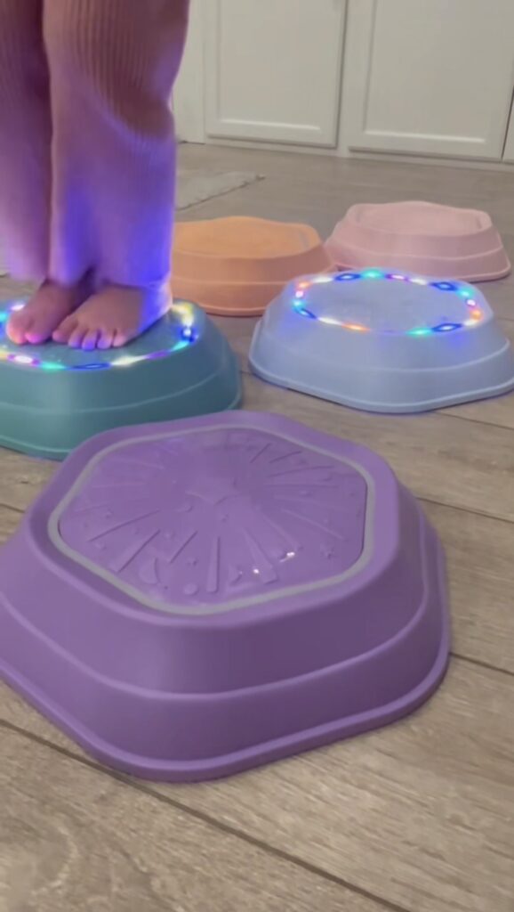 A child stands barefoot on an upturned sensory balance toy with colorful lights, surrounded by other pastel-colored bowls on a wooden floor.