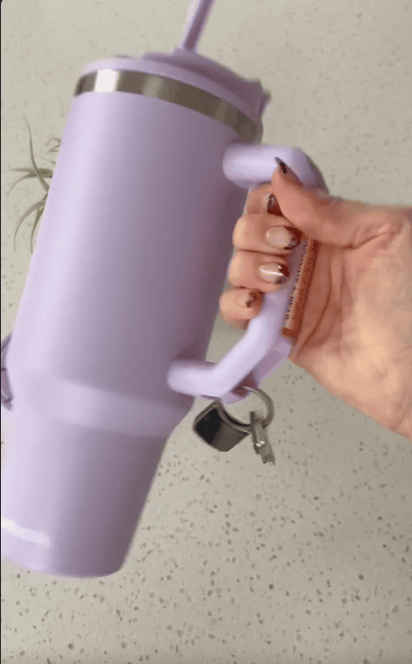 A hand with manicured nails holds a large lavender travel mug with handle and lid, complete with straw; a pair of keys hangs from the mug’s handle. A green plant is partially visible in the background.