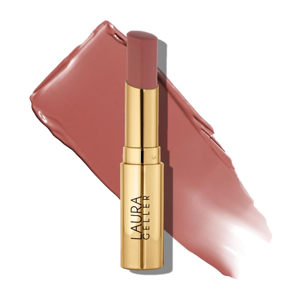 A gold lipstick tube labeled "LAURA GELLER" stands upright in front of a swatch of mauve-pink lipstick smeared diagonally in the background.