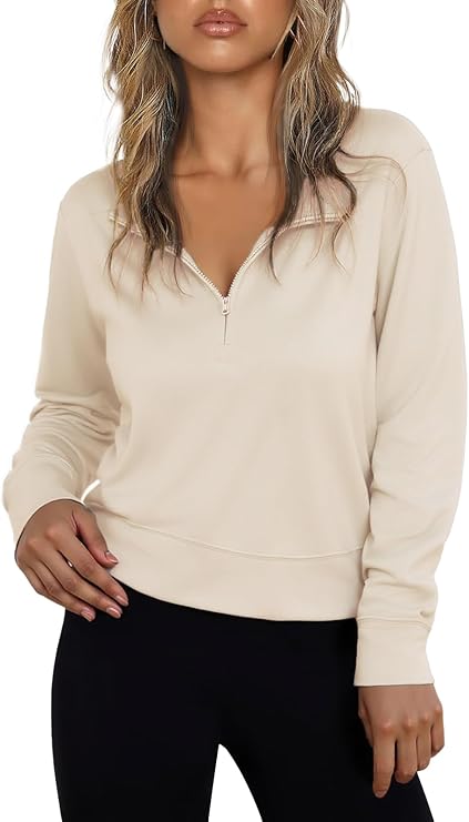 A woman wearing a light beige long-sleeve zip-up pullover with a short zipper at the neckline and black pants, standing with one hand resting near her hip.