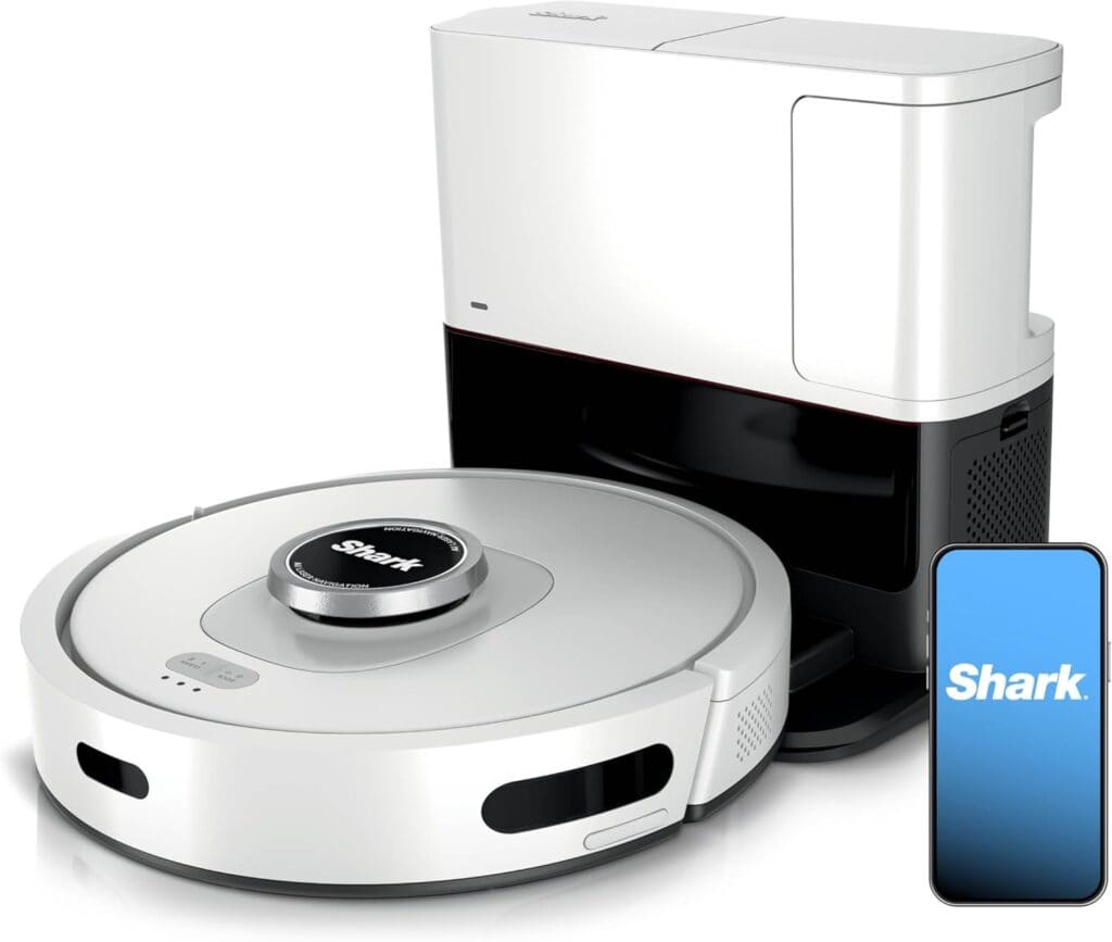 A white Shark robot vacuum with a round shape is docked at its black and white charging station. A smartphone displaying the Shark app logo is shown beside the vacuum.