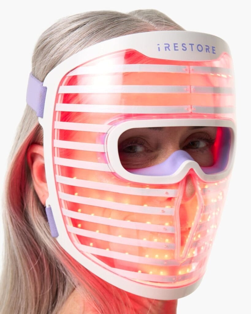 A person with long, gray hair wears an iRestore LED light therapy mask that emits a red glow, covering their entire face except for the eyes; the mask is secured with purple straps.