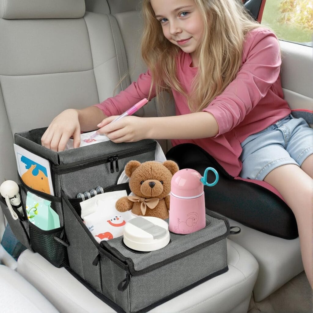 A young girl draws in the back seat of a car using a gray car organizer filled with art supplies, a teddy bear, a pink water bottle, tissues, and other items. She sits on a booster seat, smiling while drawing.