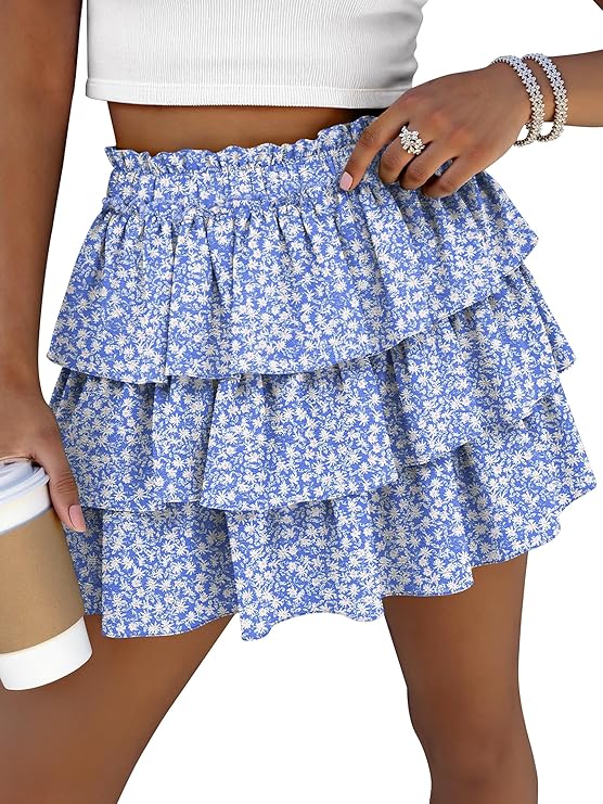 A woman wearing a white crop top and a blue floral ruffle mini skirt holds a coffee cup in one hand. She accessorizes with bracelets and a ring, and is pictured from midriff to mid-thigh.