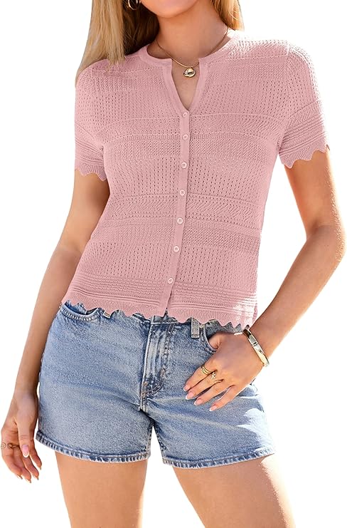 A woman wearing a light pink, short-sleeve, button-up knit top with scalloped edges, paired with light blue denim shorts and simple jewelry, stands against a plain background.