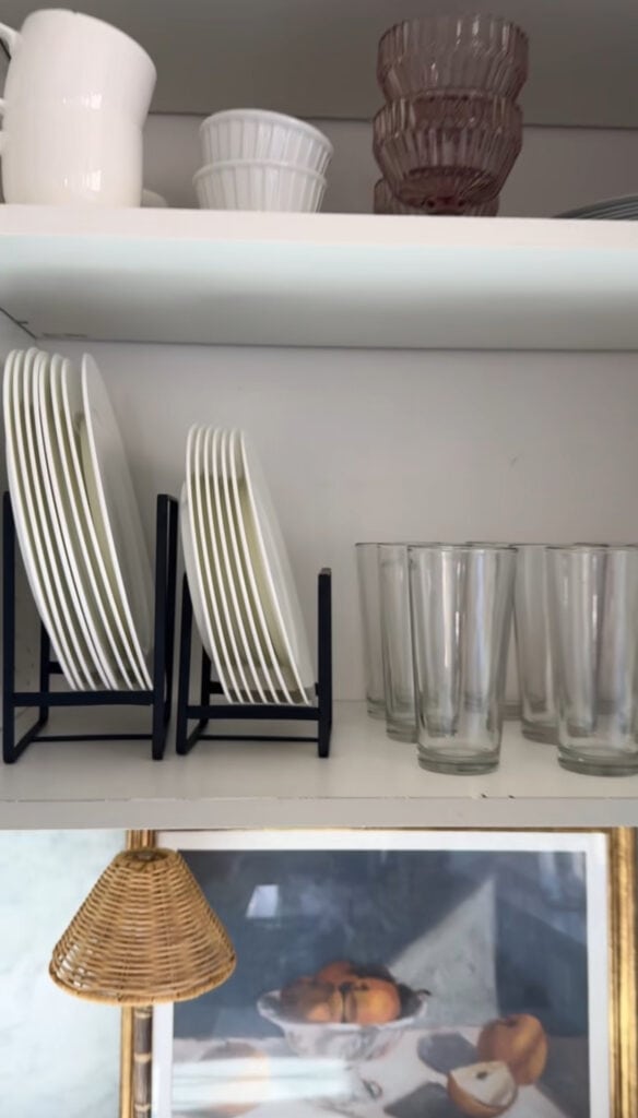 White plates in a black rack, clear glasses, and stacked bowls sit on open shelves; a reusable cake candle holder makes a thoughtful gift for moms, while a white pitcher and cups are on the top shelf beside a framed painting below.