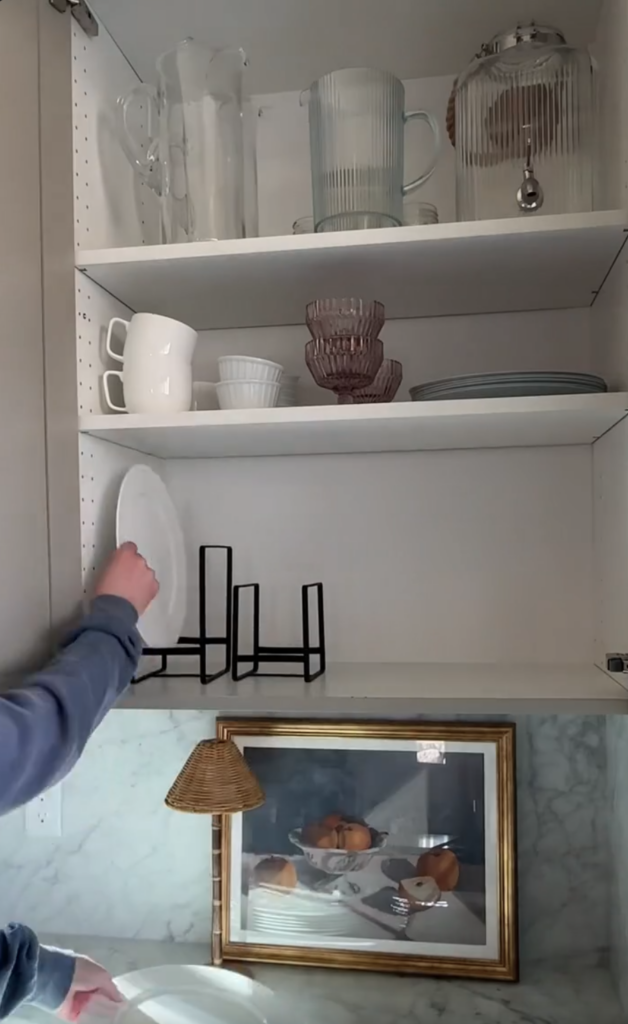 A person places a white plate onto a black plate rack inside a kitchen cabinet. The shelves above hold glass pitchers, teacups, glass bowls, and plates—perfect for moms who love to bake using their reusable cake candle holder.