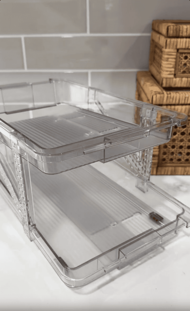 A clear, two-tier plastic storage organizer sits on a white countertop, with woven rattan boxes and a reusable cake candle holder stacked in the background against pale gray subway tile—perfect for organized moms.