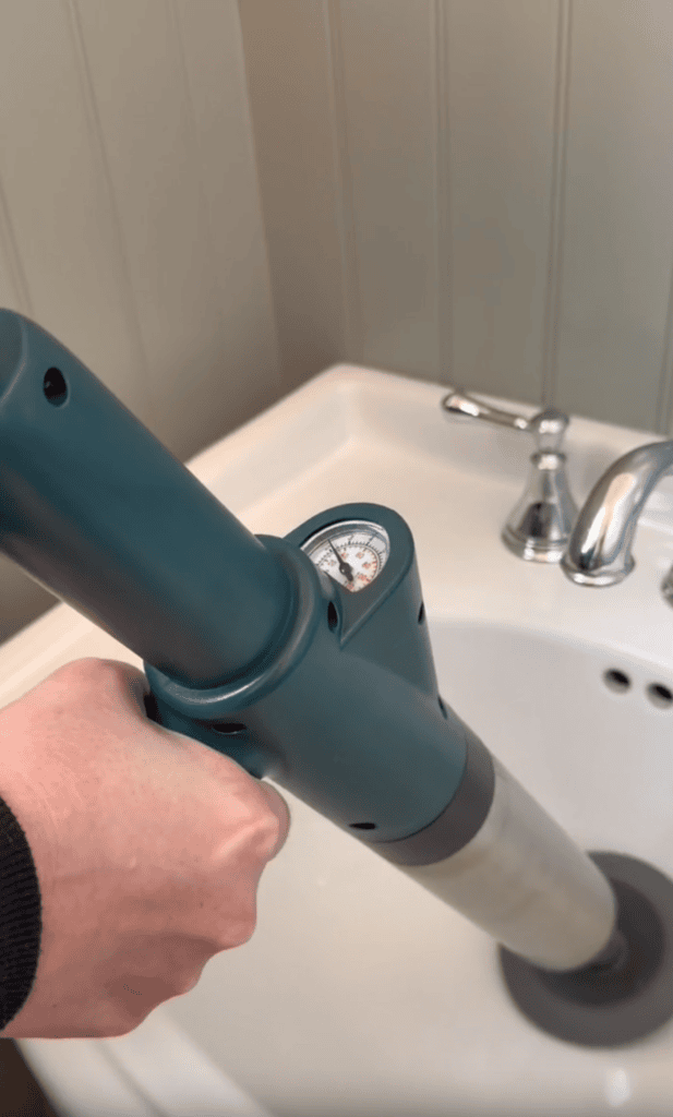 A hand holds a teal-colored plunger with a pressure gauge attached, positioned over a white bathroom sink near a silver faucet and drain—perfect for busy moms tackling everyday messes.