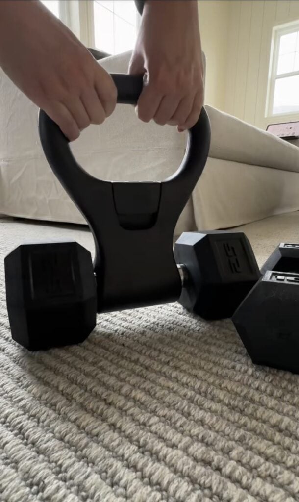 A person’s hands grip a kettlebell handle attached to a dumbbell on a carpeted floor, while nearby, two more dumbbells rest like reusable cake candle holders in a cozy, sunlit room perfect for moms' workouts.