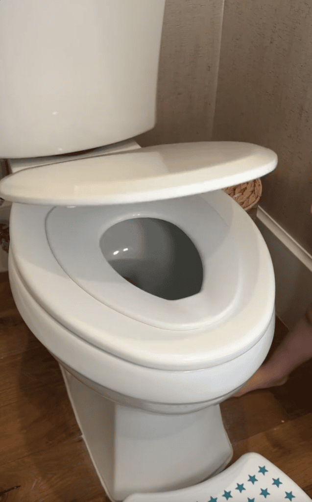 A white toilet with the lid partially closed sits on a wooden floor. A small child’s foot appears near a step stool with blue stars—a scene familiar to many moms, reminiscent of celebrating milestones like choosing the perfect reusable cake candle holder.