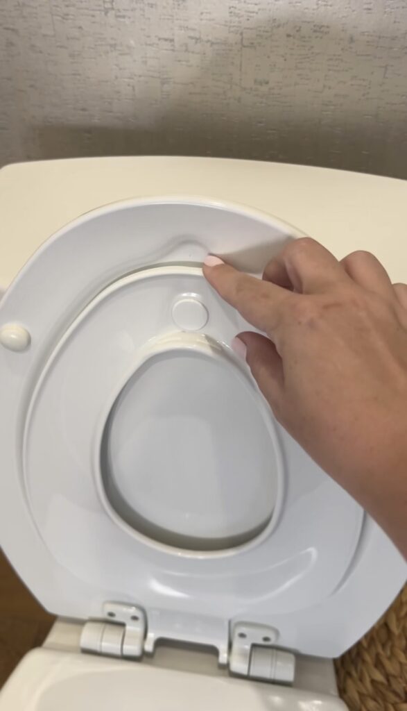 A hand with light pink nail polish presses a circular button on a white plastic toilet seat cover. A woven basket sits nearby, perfect for moms who appreciate versatile items like a reusable cake candle holder in their household.