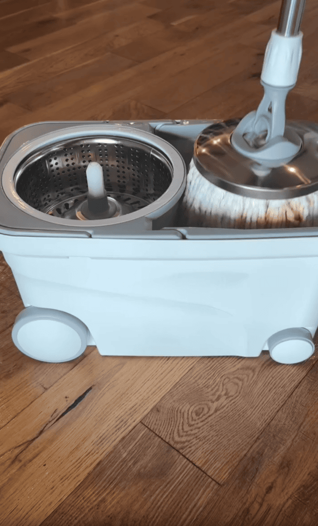 A white spin mop and bucket cleaning system with wheels sits on a wooden floor, perfect for busy moms. The mop head appears wet and is positioned above the bucket’s wringer section.