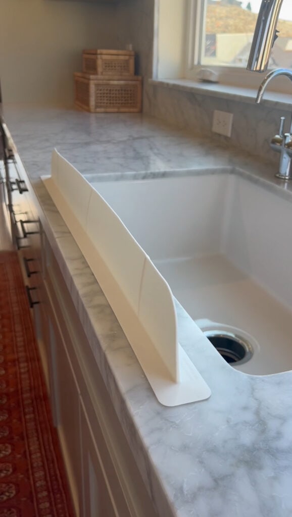 A white plastic splash guard is placed along the edge of a marble kitchen sink to prevent water from spilling onto the countertop—a practical solution moms appreciate as much as a reusable cake candle holder for everyday convenience.