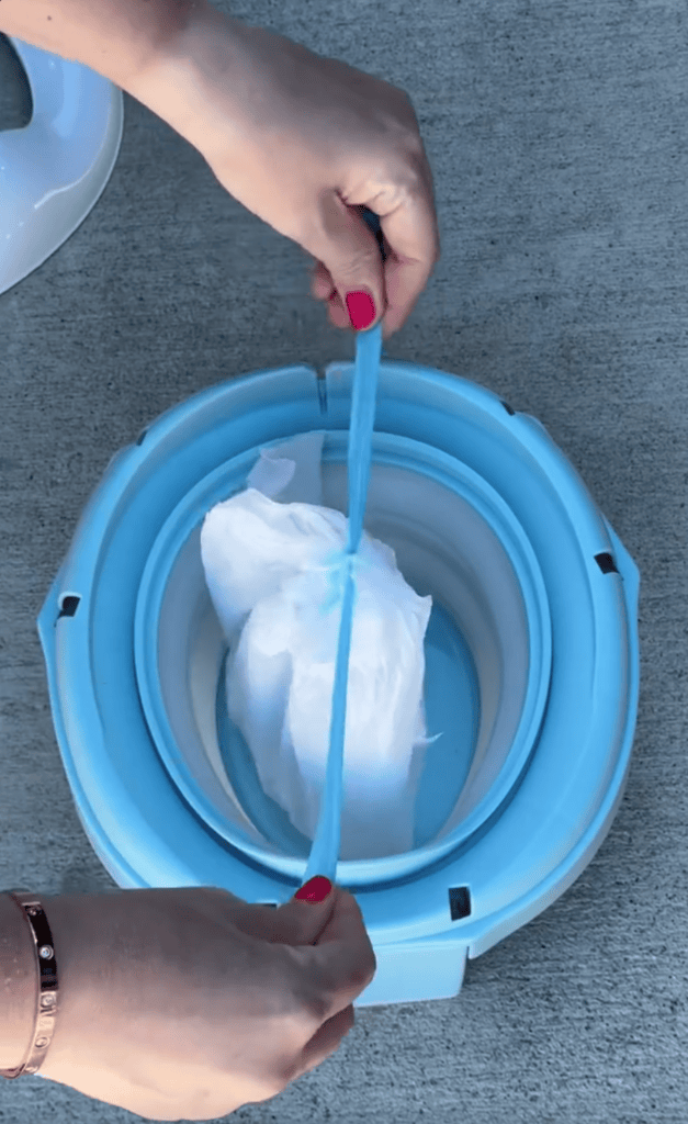 A person with red nail polish, possibly one of the busy moms, stretches a blue plastic liner over the rim of a blue portable toilet on a concrete surface—much like preparing a reusable cake candle holder for a special occasion.