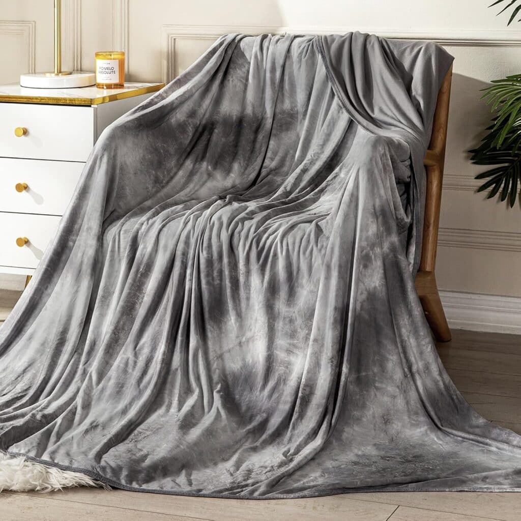 A plush, grey tie-dye blanket is draped over a wooden chair next to a white dresser with gold handles and a lit candle on top. The scene is set in a cozy, well-lit room with neutral decor.