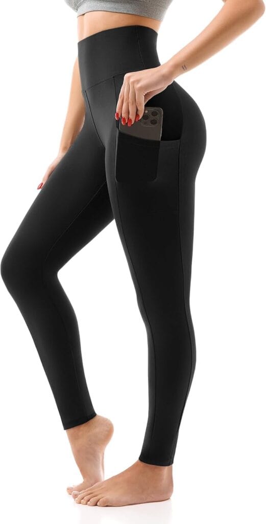 A woman wearing high-waisted black leggings with a side pocket holds a smartphone in the pocket. She is barefoot and shown from the midsection down. The leggings appear to be made of stretchy, fitted material.