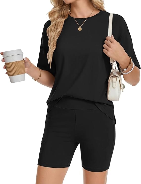 A woman wearing a black short-sleeve top and matching black shorts holds a coffee cup in one hand and a white purse over her shoulder. She wears a necklace and bracelets, with loose wavy hair.