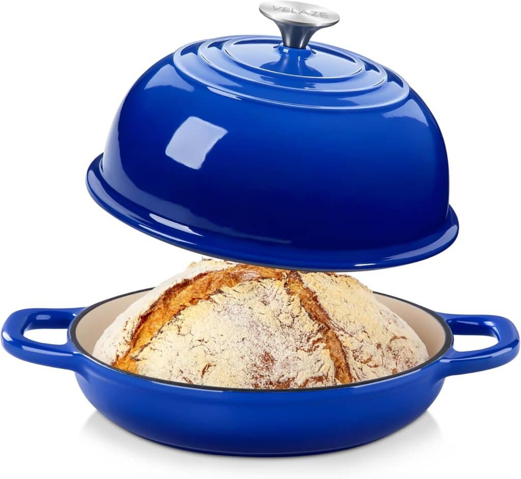 A round loaf of crusty bread sits in a blue enameled cast iron Dutch oven with handles, while the matching blue domed lid is lifted above it, revealing the fresh bread inside.