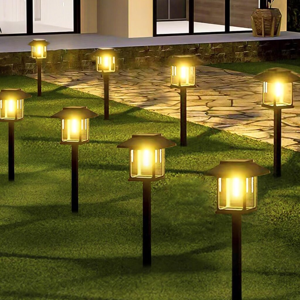A row of decorative outdoor path lights with warm yellow glow lines a stone walkway in a grassy yard outside a modern house at dusk.