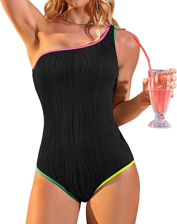 A woman wearing a black one-shoulder swimsuit with pink, green, and yellow trim holds a pink drink in a glass with a straw. Her face is not visible.