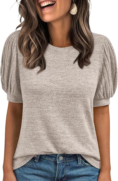 Woman wearing a light beige short-sleeve top with puffed sleeves and blue jeans, smiling with wavy brown hair and large earrings. The background is white.