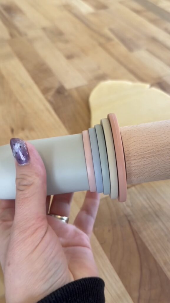 A hand grips an adjustable rolling pin with colorful rings of varying thickness; cookie dough is being evenly rolled out on a wooden surface in the background.