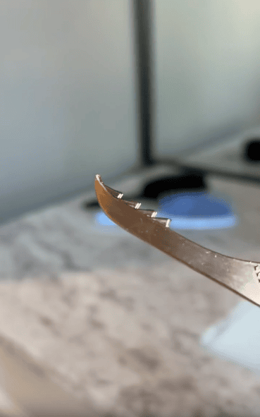 A close-up of a serrated metal tool with several small notches along its curved edge, held in front of a blurry bathroom counter and mirror in the background.