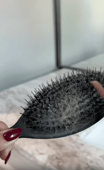 A hand with red-painted nails holds a black hairbrush filled with tangled hair against a light background with a mirror and countertop visible.