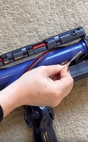 A hand uses scissors to cut hair tangled around the brush roll of a vacuum cleaner placed on a beige carpet.