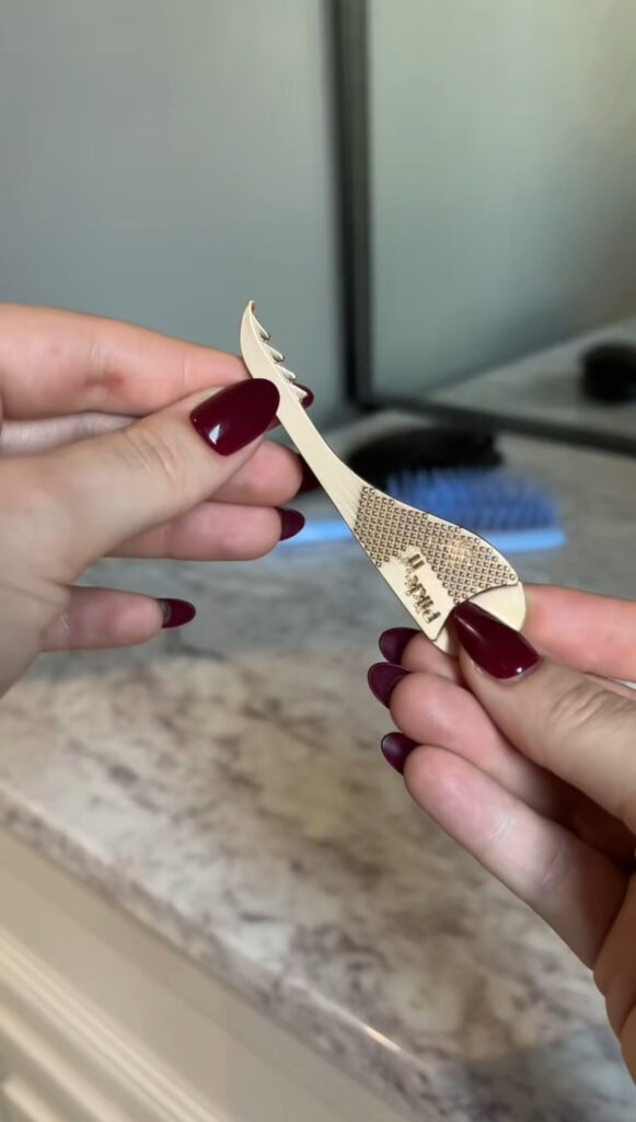 A person with dark red nail polish holds a small, metallic, textured tool with a hook and comb-like end. A hairbrush, countertop, and mirror are visible in the blurred background.