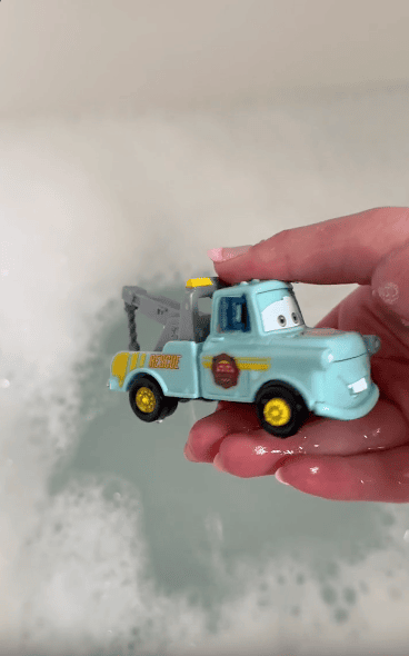 A hand holds a light blue toy tow truck with yellow details and cartoon eyes above a bathtub filled with bubbly water.