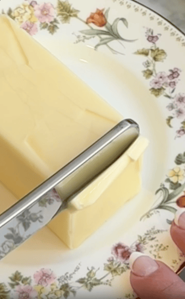 A hand holds a floral-patterned plate with a block of butter, while a knife slices a thin piece from the end.