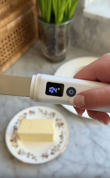 A hand holds a digital butter knife displaying a temperature of 104°F above a plate with a stick of butter on a marble countertop.