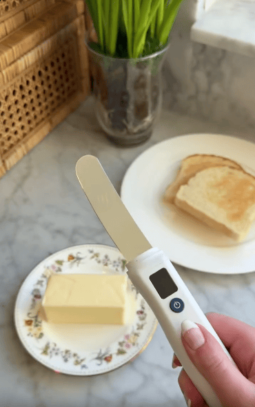 A hand holds an electric butter knife above a floral plate with a stick of butter; in the background, two slices of toast and a plant are on a marble countertop.